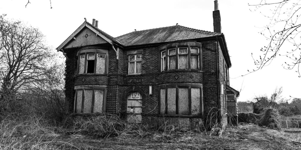 An abandoned and boarded up house. One window is open to darkness inside